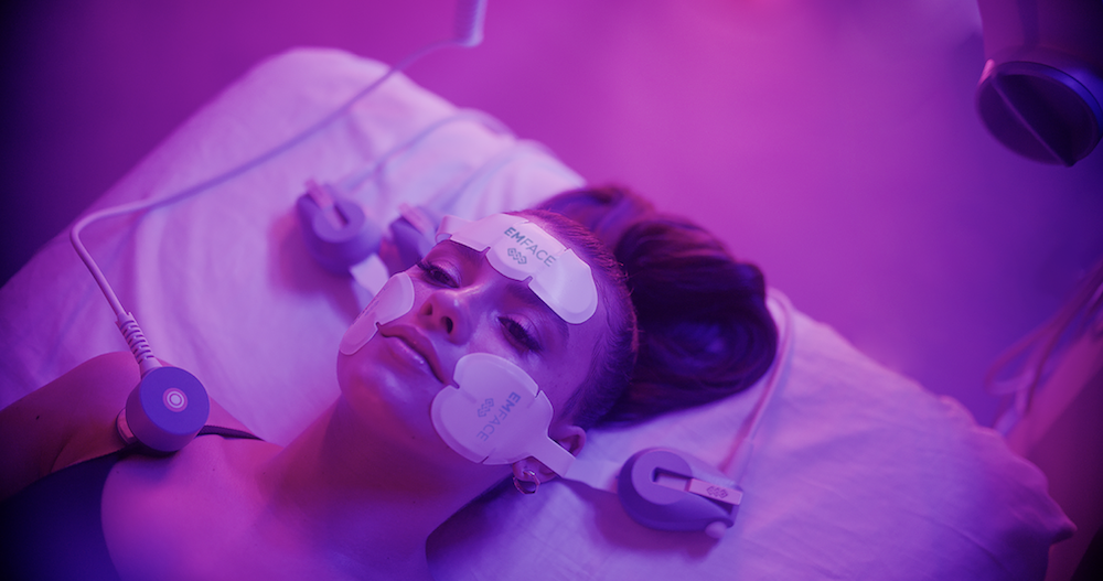 A woman receiving an Emface facial rejuvenation treatment at a medical aesthetics clinic in Wellington, FL, with applicators placed on her forehead and cheeks.